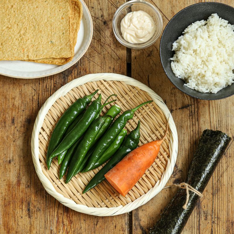 This image of the ingredients for spicy kimbap consists of green chili peppers, carrots, fish cakes, rice, and seaweed for kimbap.