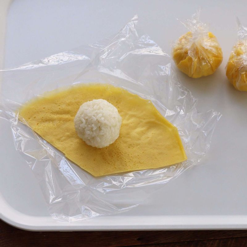 랩 위에 지단을 깔고 주먹밥을 올린 후 랩으로 동그랗게 감싸 모양을 잡아주세요. 랩을 벗겨내고 데친 미나리를 둘러 묶어주세요. (쌈무도 동일한 과정으로 동그랗게 모양을 잡아주세요)