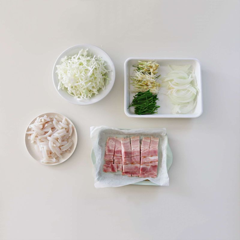 양배추는 가늘게 채를 썰어 찬물에 담갔다가 물기를 빼주세요. 달래는 3센치 길이로 썰고, 양파는 채를 썰어주세요. 오징어는 내장과 껍질을 제거해 3센치 길이로 가늘게 썰고, 베이컨도 3센치 길이로 썰어주세요.