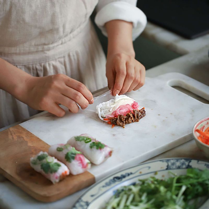 볼에 따뜻한 물을 넣고 라이스페이퍼를 5초 정도 담근 후 꺼내 평평한 접시나 도마에 라이스페이퍼를 펼치고, 절인 야채와 오이, 구운 고기, 쌀국수 면, 청양고추, 고수를 올린 후 양옆을 접어 돌돌 말아주세요.