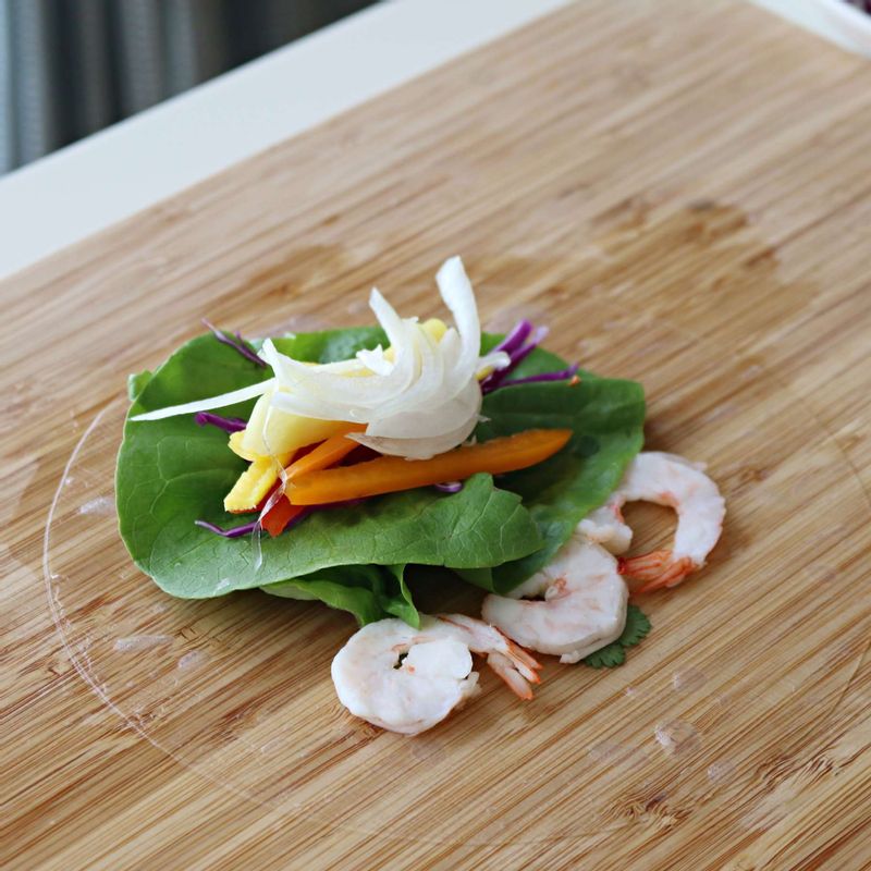 볼에 따뜻한 물을 준비한 후 라이스페이퍼를 5초정도 담근 후 꺼내주세요. 평평한 그릇이나 도마에 라이스페이퍼를 펼치고 새우와 각종 채소, 과일을 월남쌈의 1/3지점에 놓아주세요.