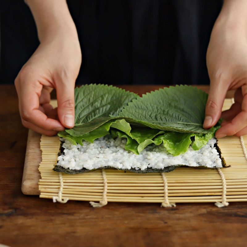 김발에 김을 올리고 밥을 고르게 편 후 청상추와 깻잎을 깔아주세요. 준비한 재료를 올려 김밥을 단단하게 말아주세요.