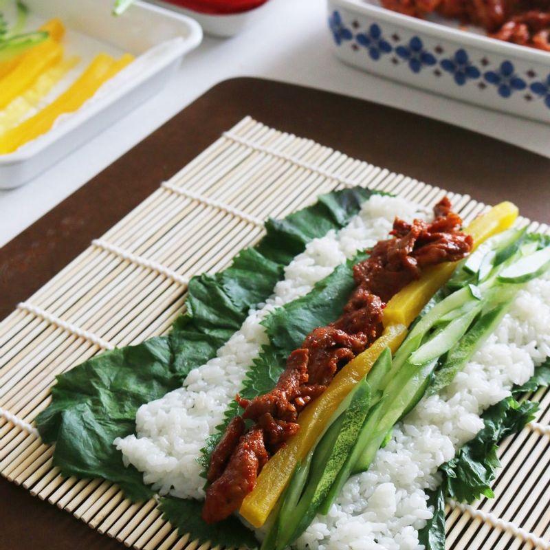 김발 위에 곰취 잎을 깔고 밥을 고르게 펴주세요. 그 위에 곰취 잎을 한 장 더 깔고 돼지고기, 오이, 단무지를 얹어 돌돌 말아줍니다.